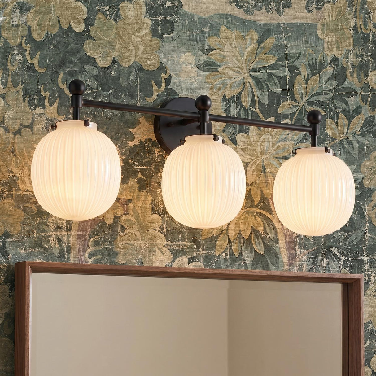 A transitional bathroom featuring the Basile 3 Light Vanity in matte black and ribbed milk glass mounted over a wood-framed mirror against green floral wallpaper.
