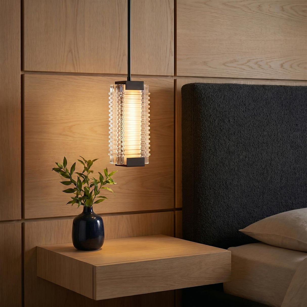 A modern bedroom with an urban bronze and clear ribbed glass pendant hanging over a floating wood nightstand beside a black textured headboard and oak wall panels.