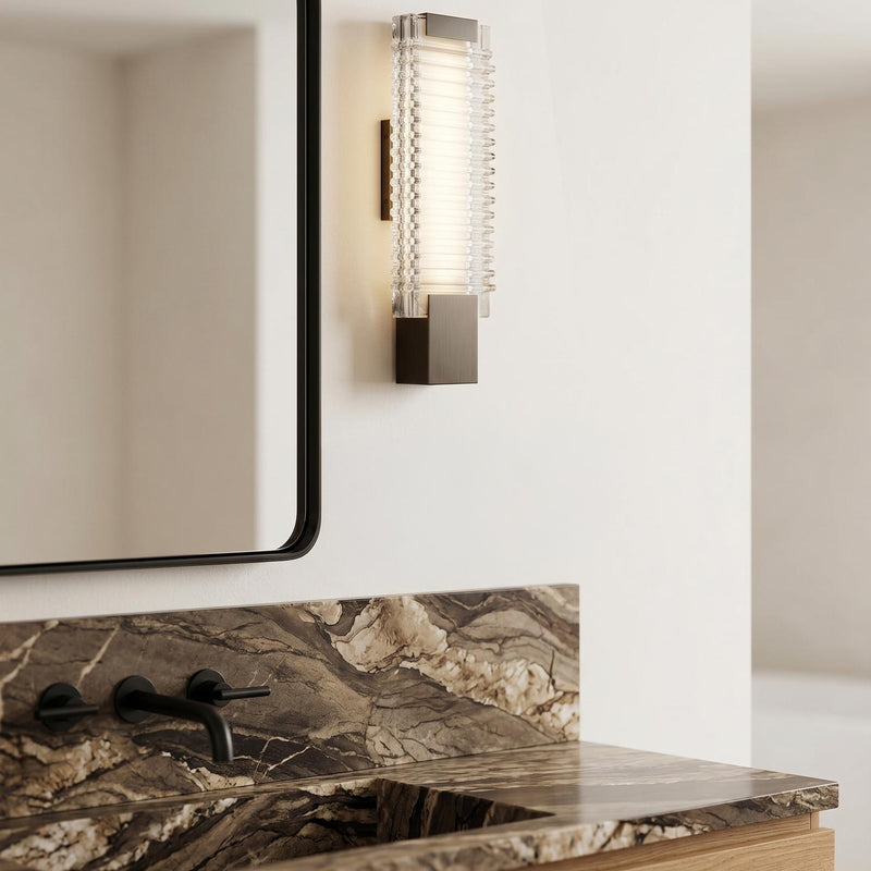 A modern bathroom with an Enders Small LED Wall Sconce in urban bronze and clear ribbed glass mounted beside a black-framed mirror above a brown marble vanity.