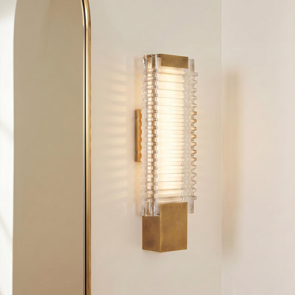 A modern bathroom features the Enders Small LED Wall Sconce in vintage brass and clear ribbed glass mounted on a white wall beside a gold arched mirror.