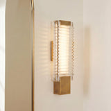 A modern bathroom features the Enders Small LED Wall Sconce in vintage brass and clear ribbed glass mounted on a white wall beside a gold arched mirror.