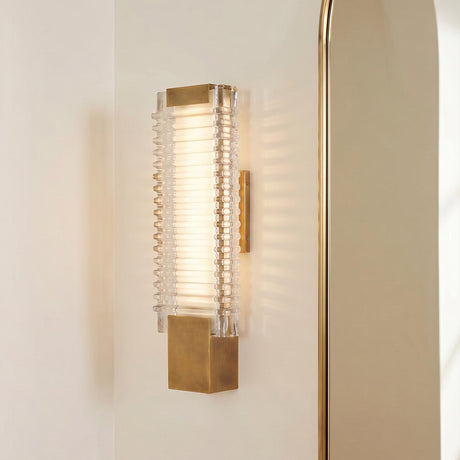 A modern bathroom features the Enders Small LED Wall Sconce in vintage brass and clear ribbed glass mounted on a white wall beside a gold arched mirror.
