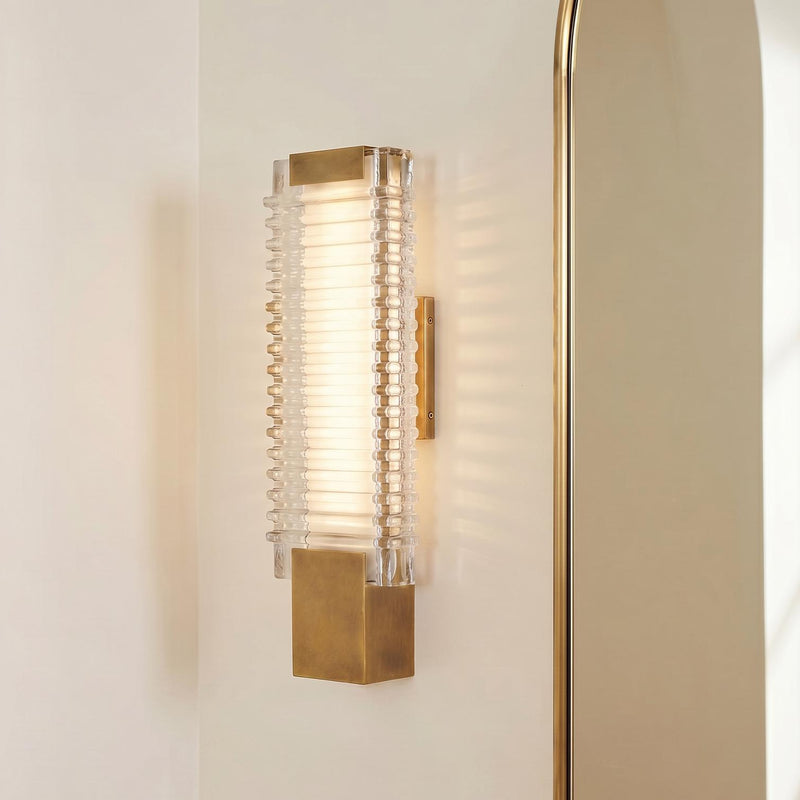A modern bathroom features the Enders Small LED Wall Sconce in vintage brass and clear ribbed glass mounted on a white wall beside a gold arched mirror.