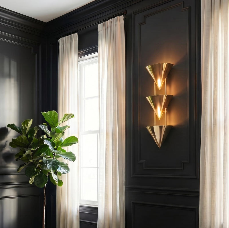 A traditional living room featuring the Zora 3 Light Wall Sconce 28", Polished Brass on charcoal paneled walls next to white linen curtains and a fiddle leaf fig.