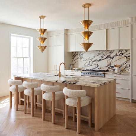 A contemporary kitchen with two Zora 3 Light Pendant 10", Polished Brass fixtures over a marble island with boucle bar stools and a bold marble backsplash.