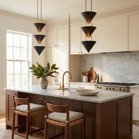 A contemporary kitchen featuring two Zora 3 Light Pendants in urban bronze and polished brass finishes over a white marble island with dark wood cabinets and woven stools.