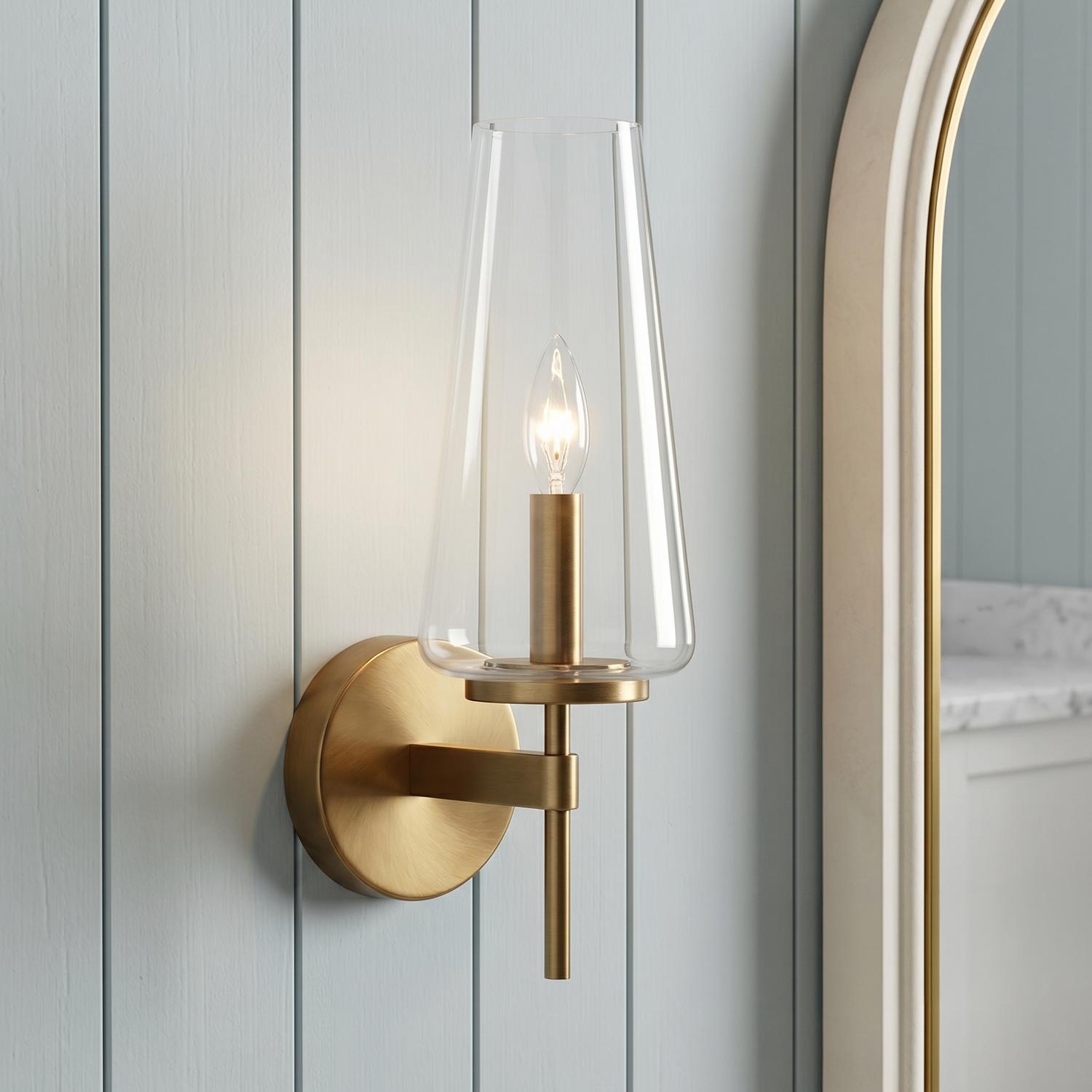 A coastal bathroom featuring the Darlene Wall Sconce in aged brass with a tapered clear glass shade mounted on light blue shiplap walls beside an arched mirror.