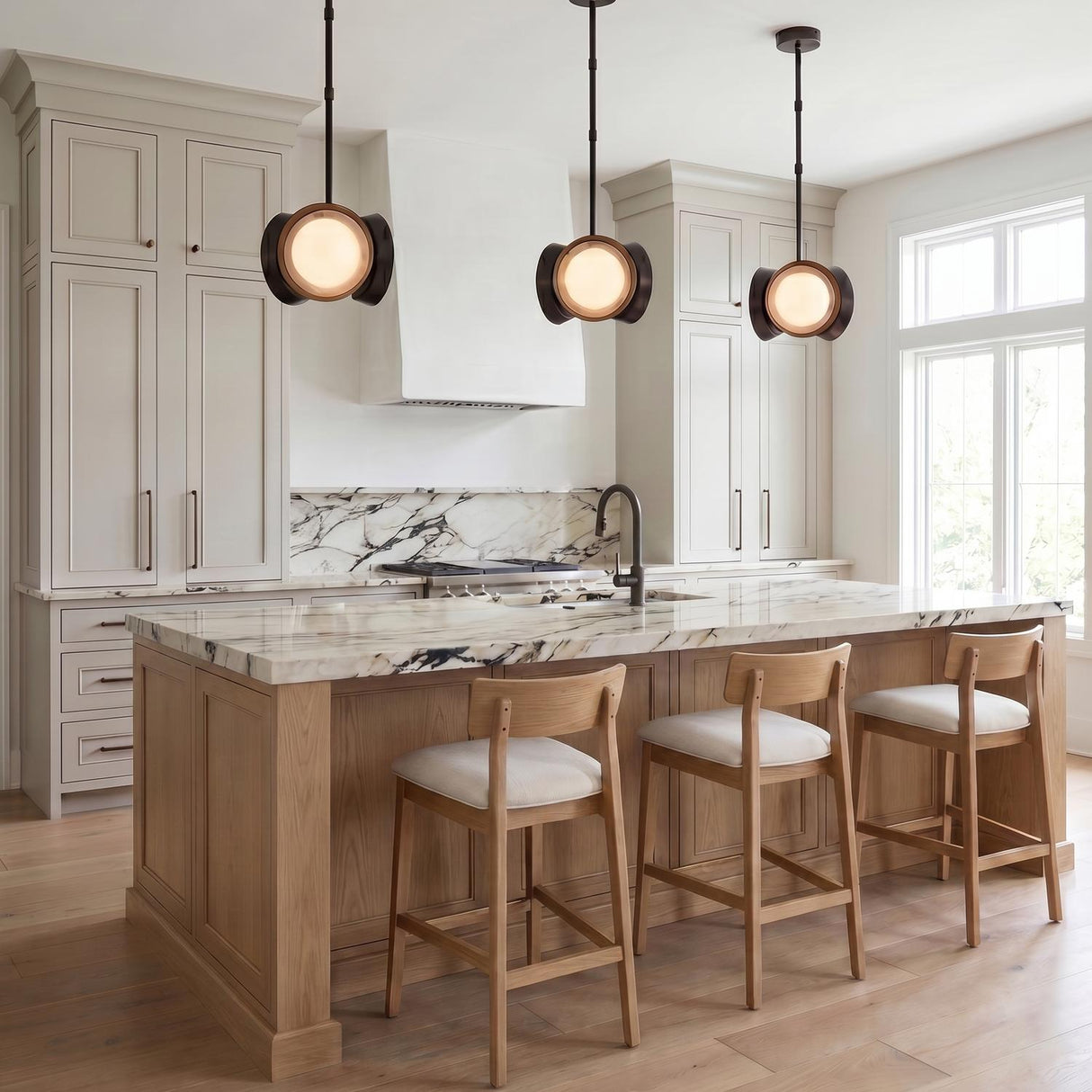 A contemporary kitchen with three Osmund LED Pendants in urban bronze over a white marble island with light wood bar stools and off-white shaker cabinetry.