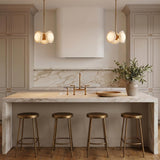 A contemporary kitchen with two Osmund LED pendants in vintage brass and alabaster over a white marble waterfall island featuring brass stools and beige cabinets.