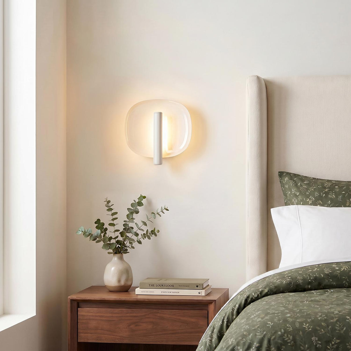 A modern bedroom with a pearl white Orlene LED Wall Sconce mounted above a walnut nightstand beside a cream upholstered headboard and green bedding.