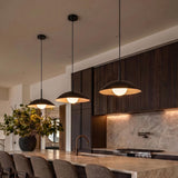 A modern kitchen with three matte black and frosted glass saucer pendants hanging over a marble island featuring a bronze faucet and dark slatted wood cabinets.