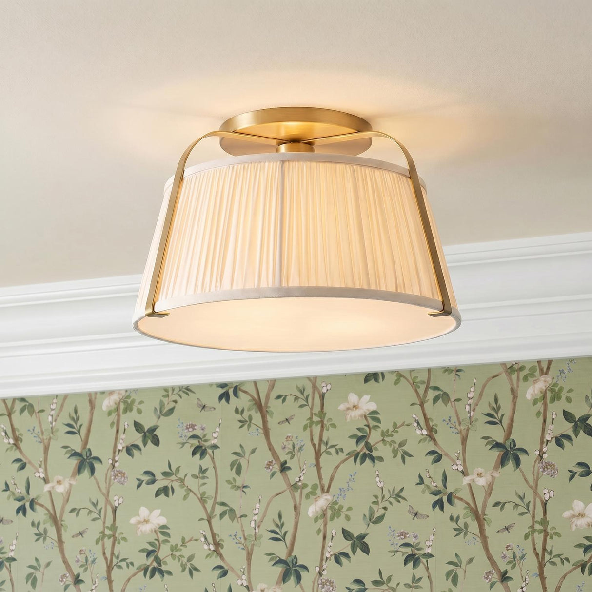 A traditional bedroom with a brushed brass semi-flush mount and pleated linen shade installed on a standard ceiling above sage green floral wallpaper and white molding.