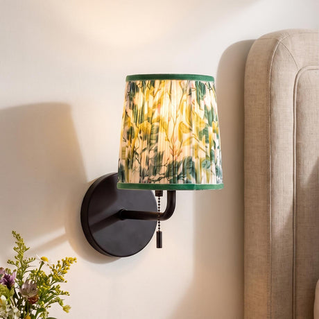 A transitional bedroom features the Daina Wall Sconce 10", Matte Black and Green Floral with a pleated fabric shade mounted beside a beige upholstered headboard.