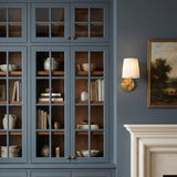 A traditional living room with the Daina Wall Sconce 10", Brushed Brass and White mounted on a blue wall beside glass-front cabinets with walnut interiors.