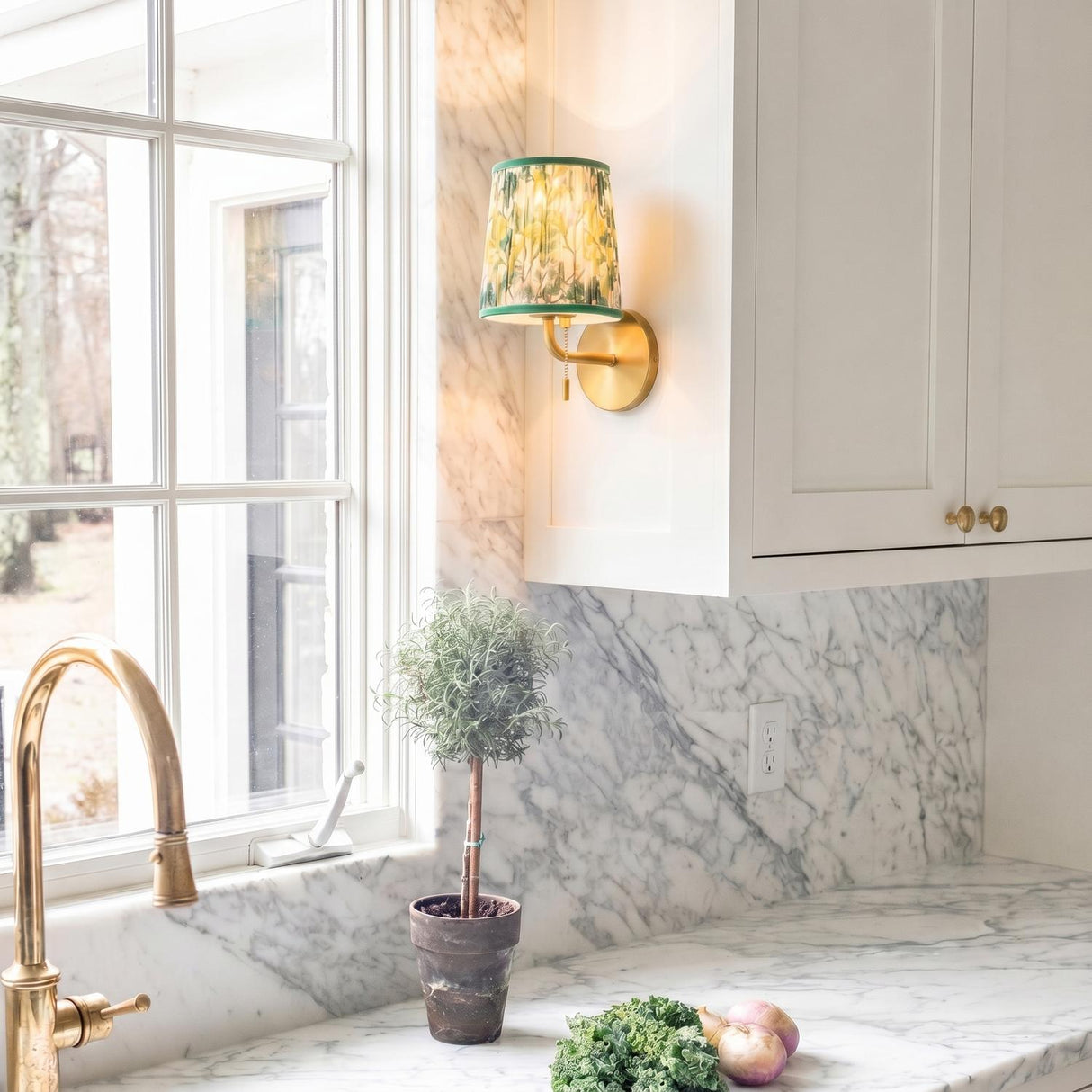 A transitional kitchen with the Daina Wall Sconce in brushed brass and green floral mounted beside a window above a marble backsplash and white cabinets.
