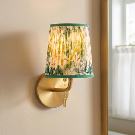 A traditional bedroom features a Daina Wall Sconce 10 inch Brushed Brass and Green Floral with a pleated shade and pull chain mounted beside a gold mirror.