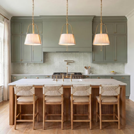 A transitional kitchen with three brushed brass pendants and white linen shades suspended over a white marble island and sage green cabinetry with tile backsplash.