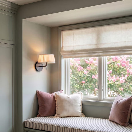 A transitional bedroom nook with a Daina Wall Sconce 7" in matte black with a white pleated shade mounted beside a striped cushion window seat and pink pillows.