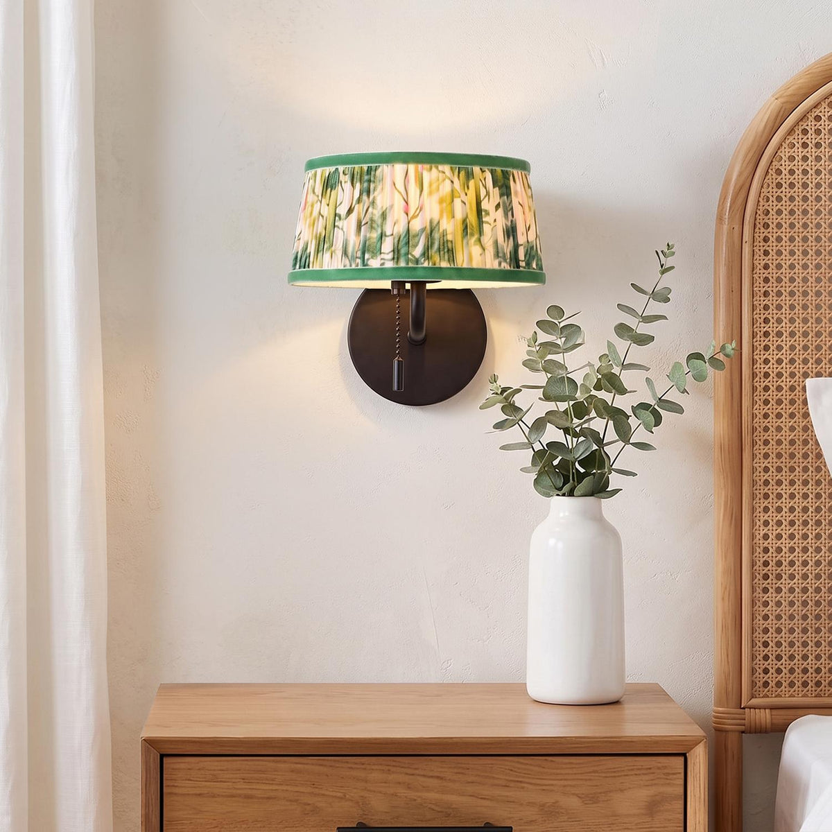 A cottage bedroom with the Daina Wall Sconce 7", Matte Black and Green Floral, installed above a natural wood nightstand next to a rattan headboard.