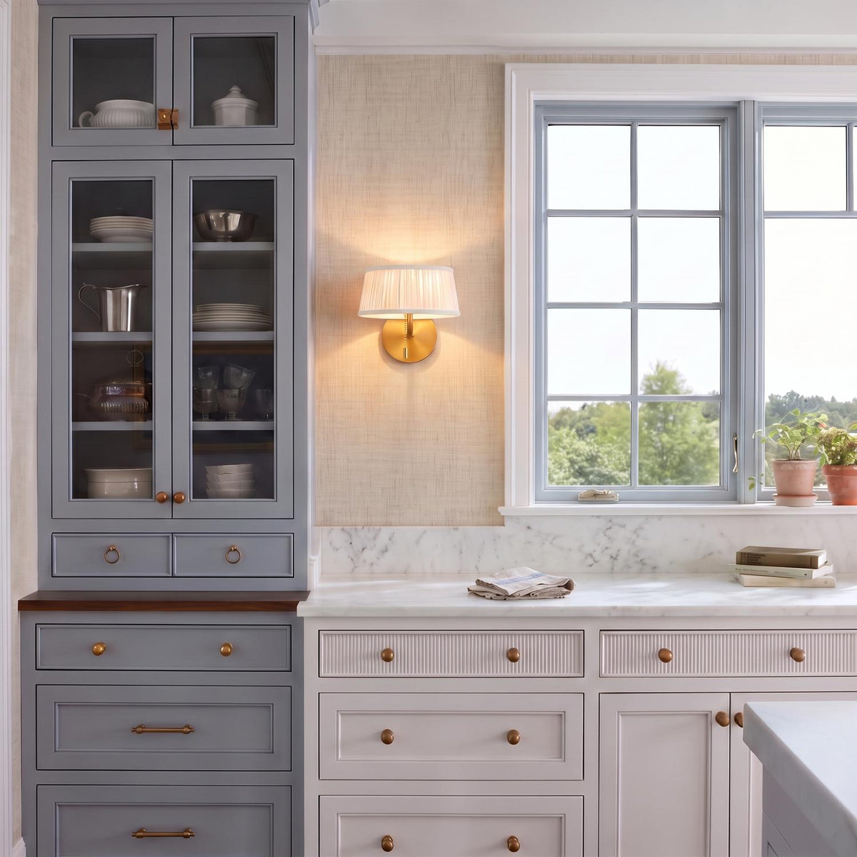 A transitional kitchen with the Daina Wall Sconce 7", Brushed Brass and White mounted on beige wallpaper between gray cabinetry and a window above marble countertops.