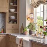 A traditional kitchen with the Daina Wall Sconce in brushed brass and blue floral mounted beside a window over a marble countertop and apron sink.
