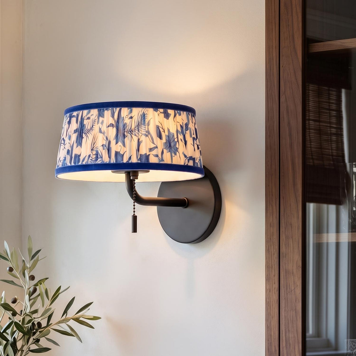 A transitional living room featuring a matte black wall sconce with a blue floral pleated fabric shade mounted along a white wall beside a walnut cabinet.