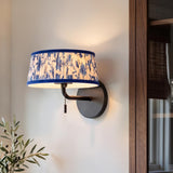 A transitional living room featuring a matte black wall sconce with a blue floral pleated fabric shade mounted along a white wall beside a walnut cabinet.