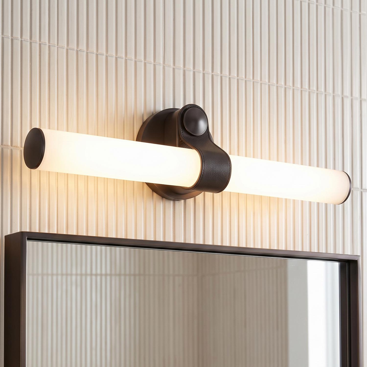 A modern bathroom features the Bodhi LED Wall Light in urban bronze and frosted glass mounted above a dark-framed mirror against vertical white kit-kat wall tiles.
