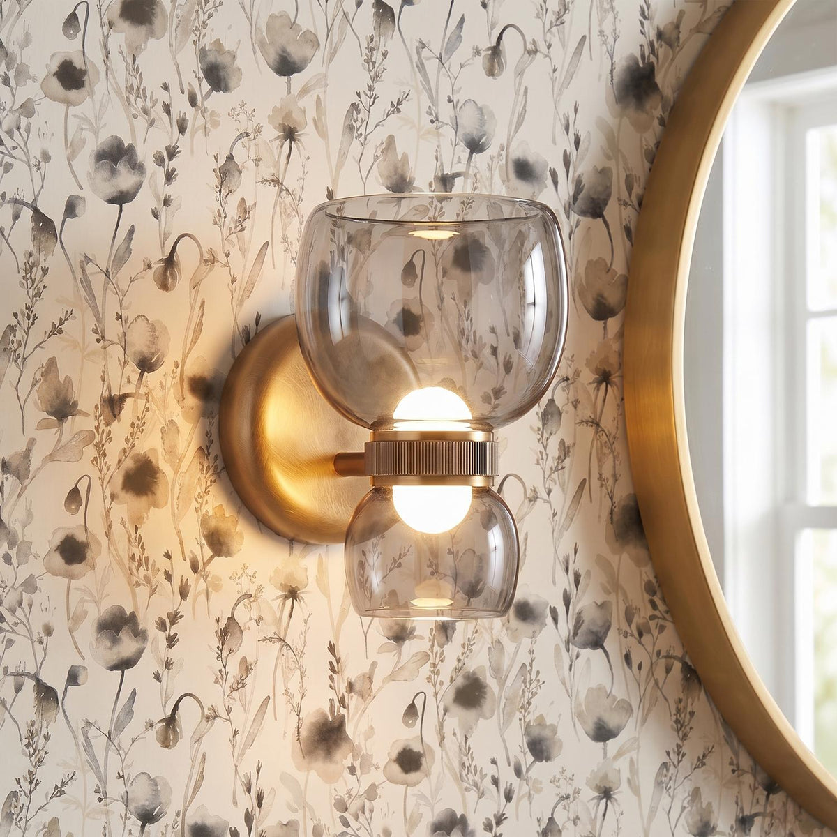 A transitional bathroom features the Brann LED Wall Sconce in vintage brass with smoke glass shades mounted on botanical wallpaper beside a round brass mirror.