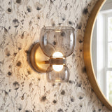 A transitional bathroom features the Brann LED Wall Sconce in vintage brass with smoke glass shades mounted on botanical wallpaper beside a round brass mirror.