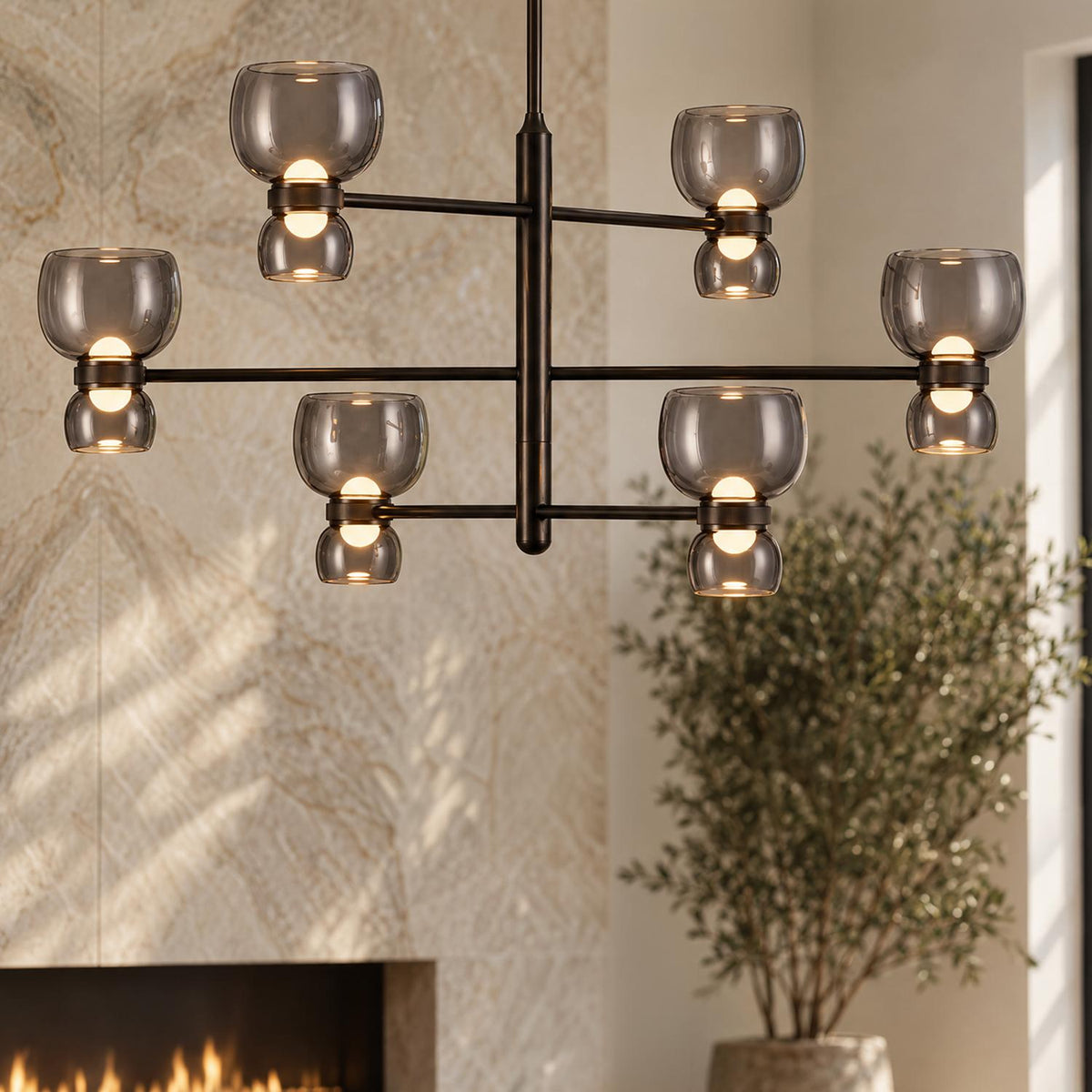 A contemporary living room featuring a Brann LED Chandelier in urban bronze with smoke glass shades suspended near a stone fireplace and potted plant.