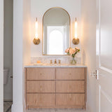 A transitional bathroom with two Videl LED Wall Sconces in vintage brass flanking an arched gold mirror over a marble countertop and wood vanity.