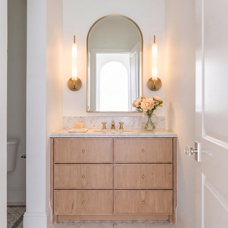 A transitional bathroom with two Videl LED Wall Sconces in vintage brass flanking an arched gold mirror over a marble countertop and wood vanity.