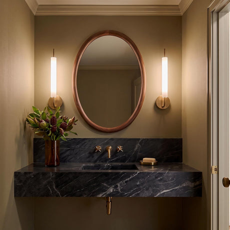 A transitional bathroom with two Videl LED Wall Sconce 20" Vintage Brass fixtures flanking an oval wood mirror above a dark marble floating vanity with brass hardware.