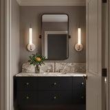 A modern bathroom with two Videl LED Wall Sconce 20", Vintage Brass and Travertine fixtures flanking a rounded mirror above a marble countertop vanity.
