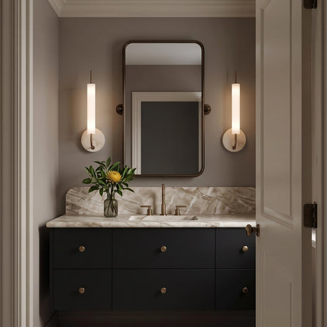 A modern bathroom with two Videl LED Wall Sconce 20", Vintage Brass and Travertine fixtures flanking a rounded mirror above a marble countertop vanity.