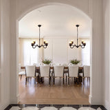 A transitional dining room with two Rubina 4 Light Chandeliers in matte black over a long wood dining table, featuring white upholstered chairs and light oak flooring.