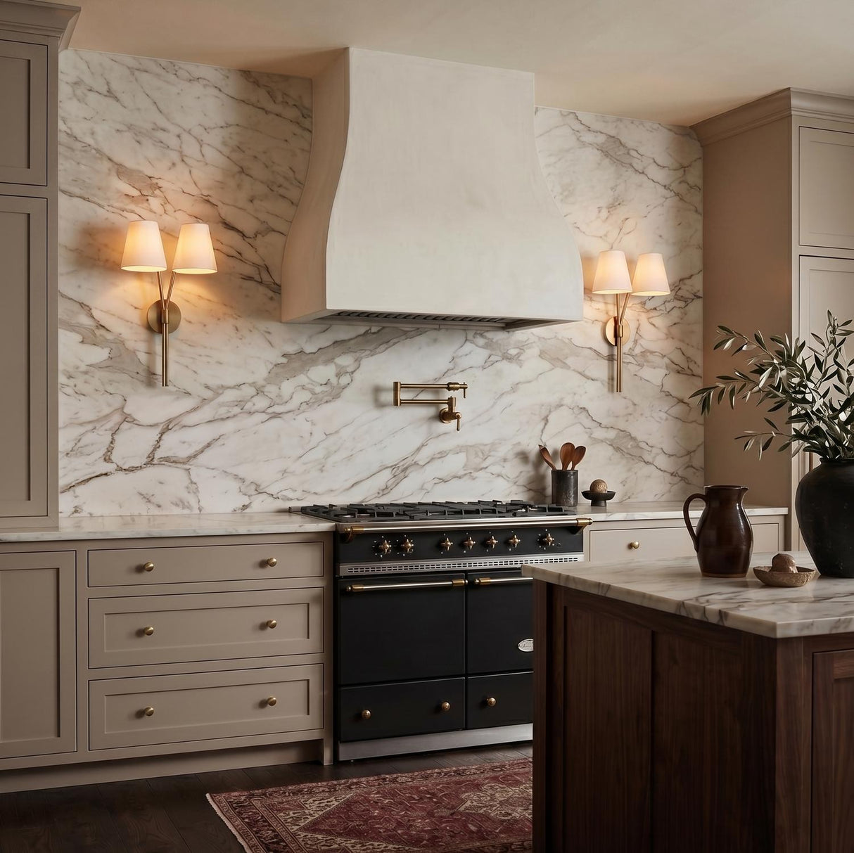 A transitional kitchen with two Althea Double Wall Sconce 23", Aged Brass fixtures featuring linen shades flanking a white plaster hood and marble backsplash.