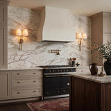 A transitional kitchen with two Althea Double Wall Sconce 23", Aged Brass fixtures featuring linen shades flanking a white plaster hood and marble backsplash.