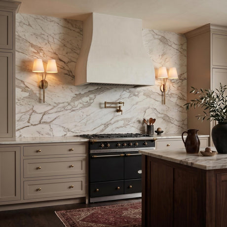 A transitional kitchen with two Althea Double Wall Sconce 23", Aged Brass fixtures featuring linen shades flanking a white plaster hood and marble backsplash.