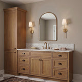 A transitional bathroom with two aged brass Althea wall sconces with linen shades flanking an arched gold mirror above a wood vanity and marble countertop.