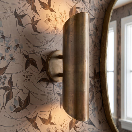 A transitional bathroom with the Kitt 2 Light Wall Sconce 16" in oxidized brass mounted on floral wallpaper beside a gold framed mirror and white window trim.