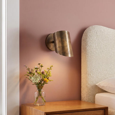 A contemporary bedroom with an oxidized brass wall sconce mounted on a mauve wall beside a white bouclé headboard and a wooden nightstand with flowers.