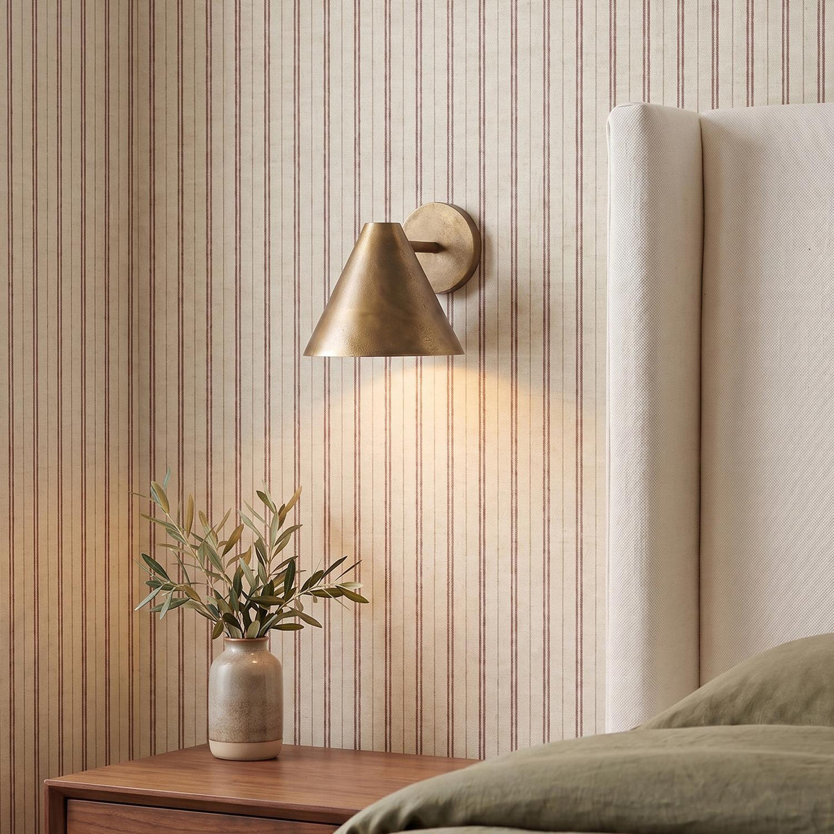 A transitional bedroom with a Kate Wall Sconce 8", Oxidized Brass, mounted against red striped wallpaper beside a white upholstered headboard and wood nightstand.