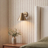 A transitional bedroom with a Kate Wall Sconce 8", Oxidized Brass, mounted against red striped wallpaper beside a white upholstered headboard and wood nightstand.