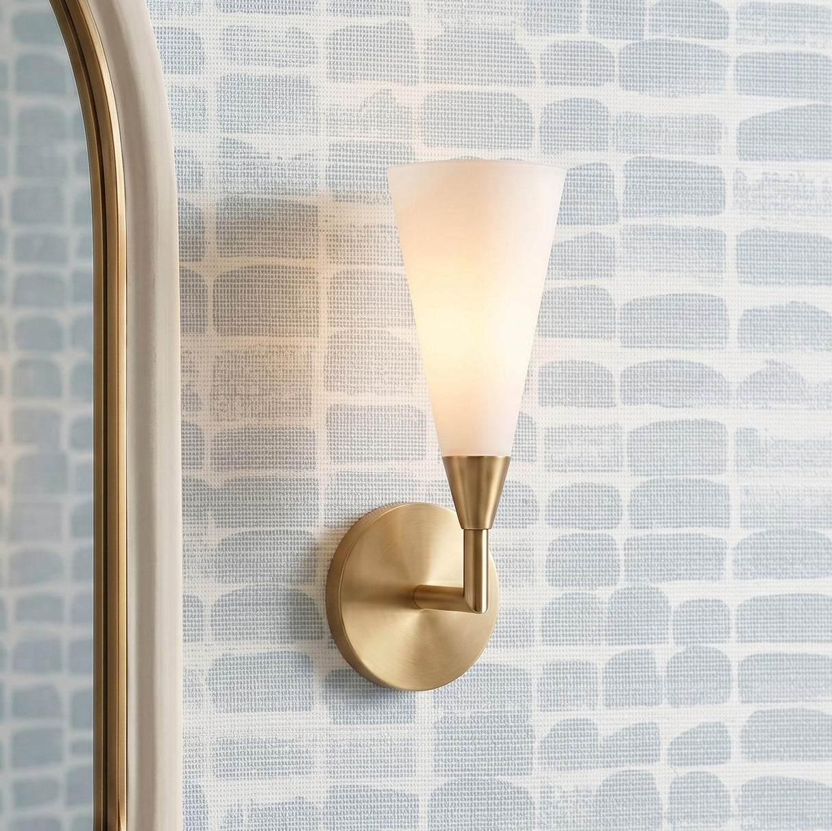 A contemporary bathroom featuring an Avani Wall Sconce 14", Matte Brass and Opal Glass mounted beside a gold arched mirror against blue and white patterned wallpaper.
