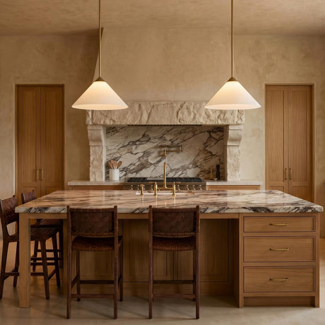 A Mediterranean kitchen with two matte brass and opal glass pendants suspended over a marble island featuring leather woven stools and a stone vent hood.