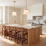 A transitional kitchen with two Camron Pendants in aged brass and opal glass over a marble-topped wood island with woven bar stools and matching marble backsplash.