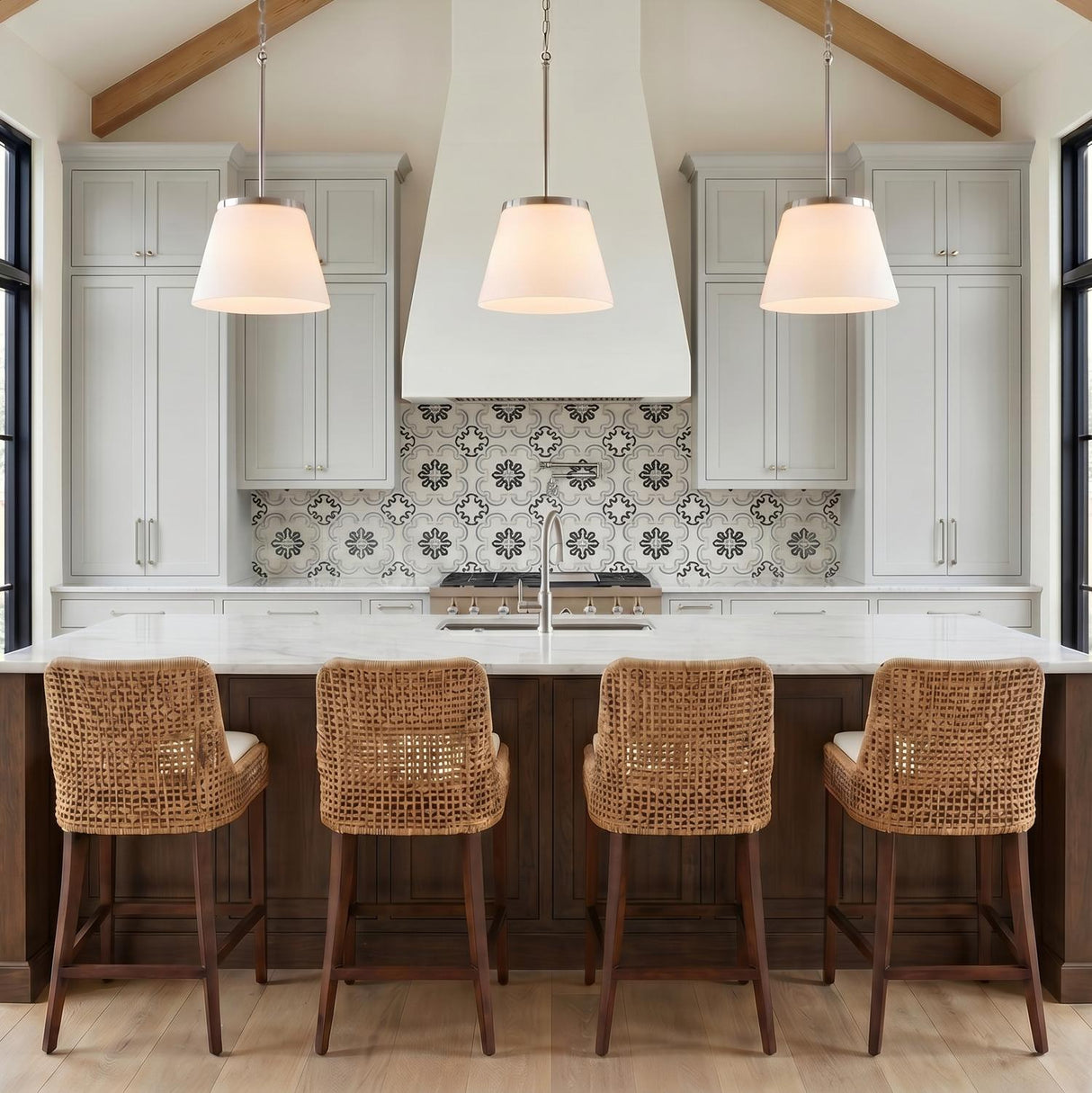 A modern farmhouse kitchen with three brushed nickel and opal glass pendants suspended over a white marble island under a vaulted beamed ceiling with woven stools.