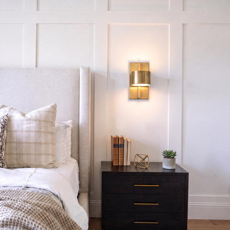 A transitional bedroom features an aged brass wall sconce mounted on a white board and batten wall beside an upholstered headboard and dark nightstand.